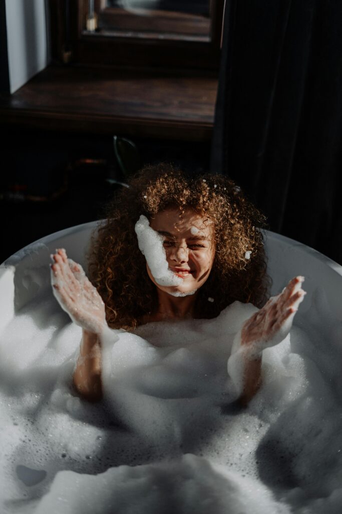 pexels-photo-4155487-4155487 A smiling young woman with curly hair enjoys a relaxing bubble bath in a cozy bathroom setting.