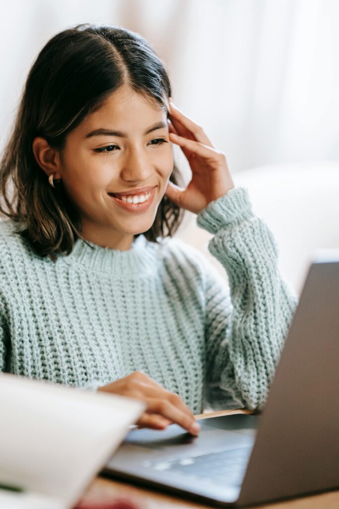 pexels-photo-6347901-6347901 A young woman smiling as she uses a laptop in a cozy indoor setting, creating a comfortable workspace atmosphere.